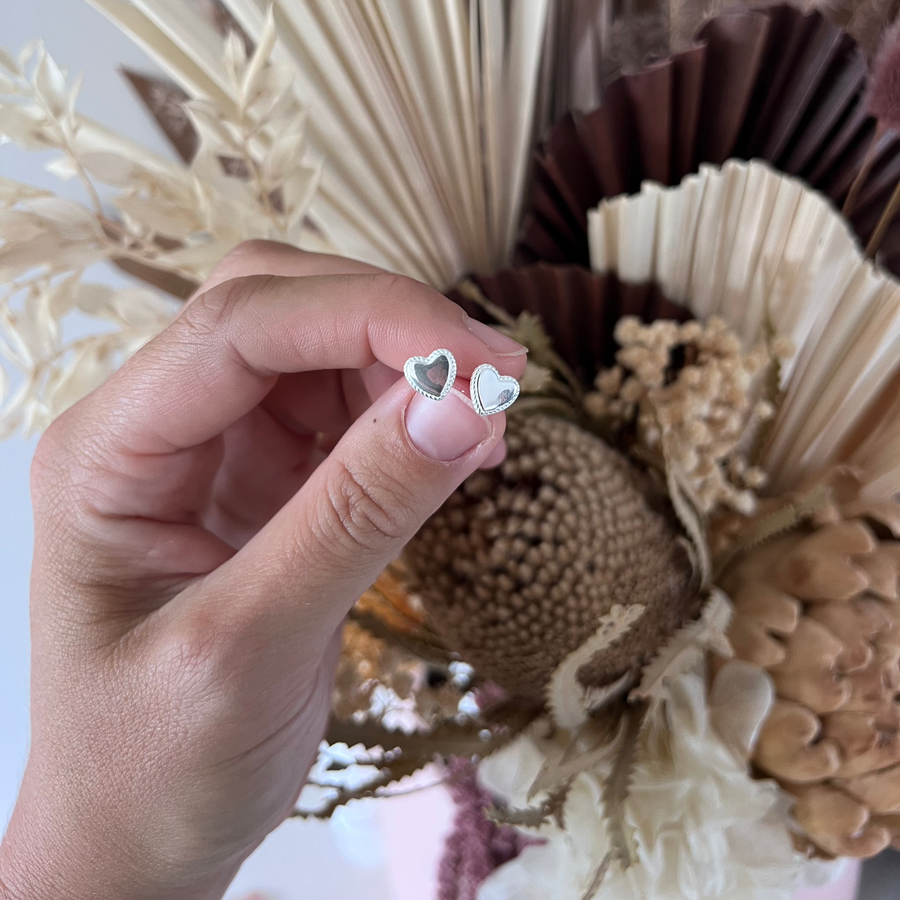 A hand holding a silver stud earring shaped like a heart, with a decorative flower arrangement in the background.