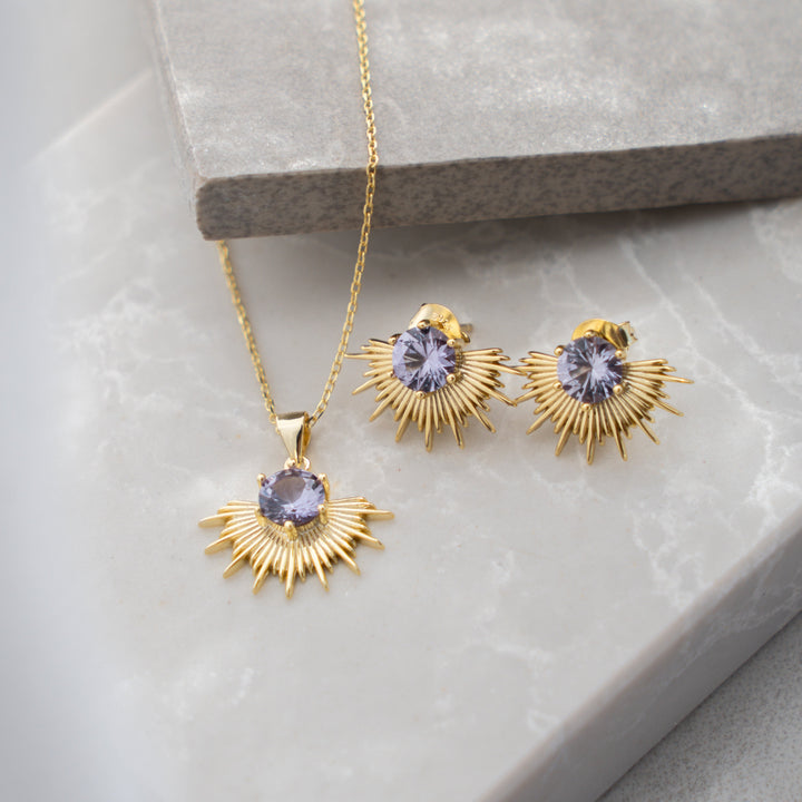 Gold jewelry set with sunburst design and purple gemstones on a light gray surface.