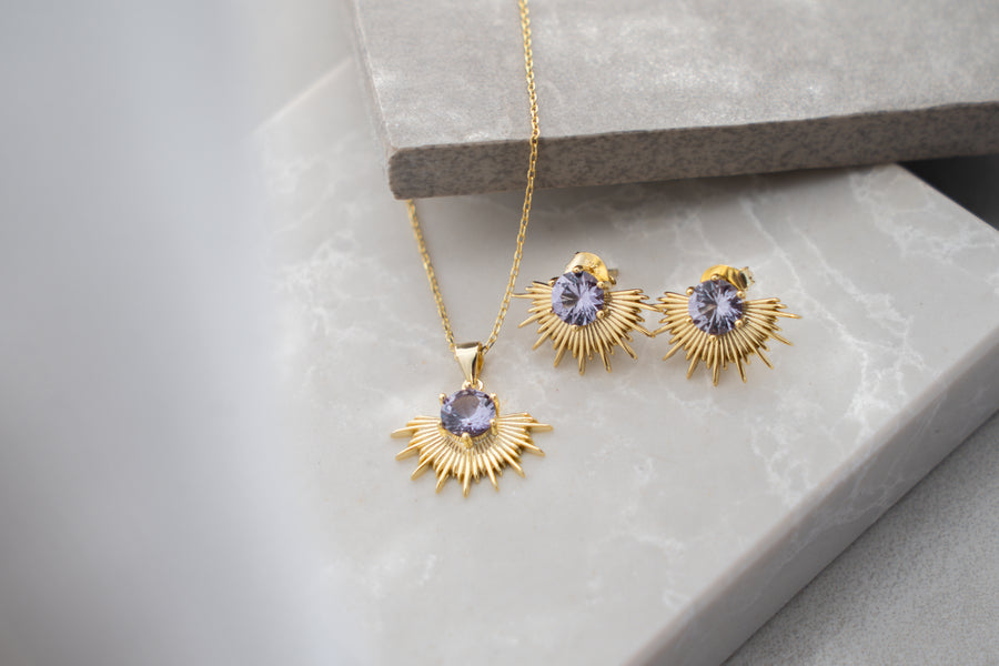 Gold jewelry set with sunburst design and purple gemstones on a light gray surface.
